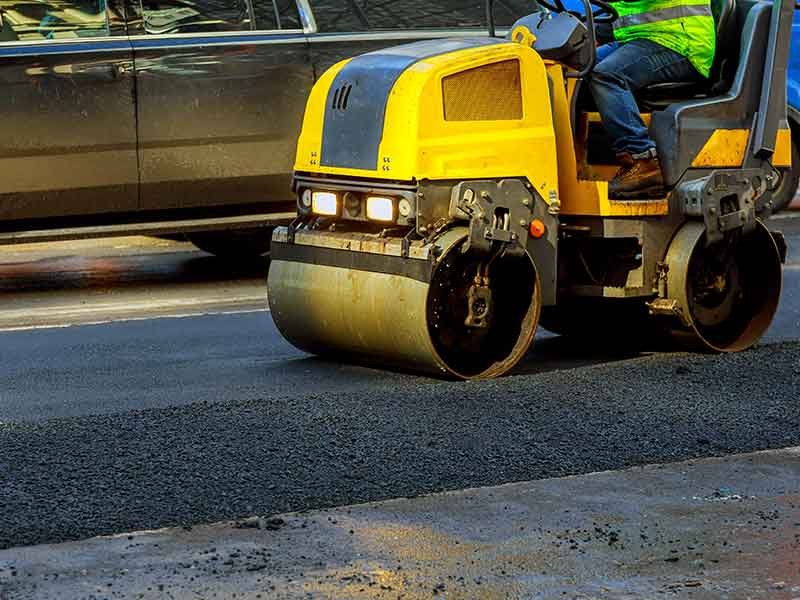 Aman driving yellow roller on paving, wearing safety jacket.