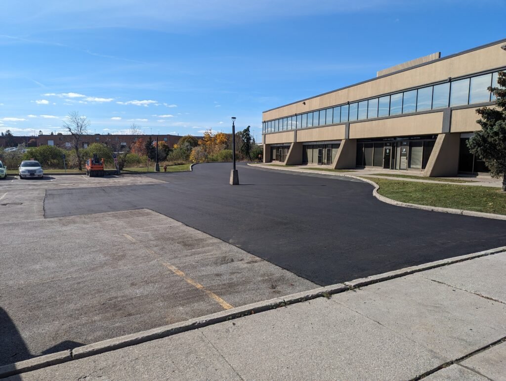Newly paved section of a parking lot with a lamp post and office building on a clear day.