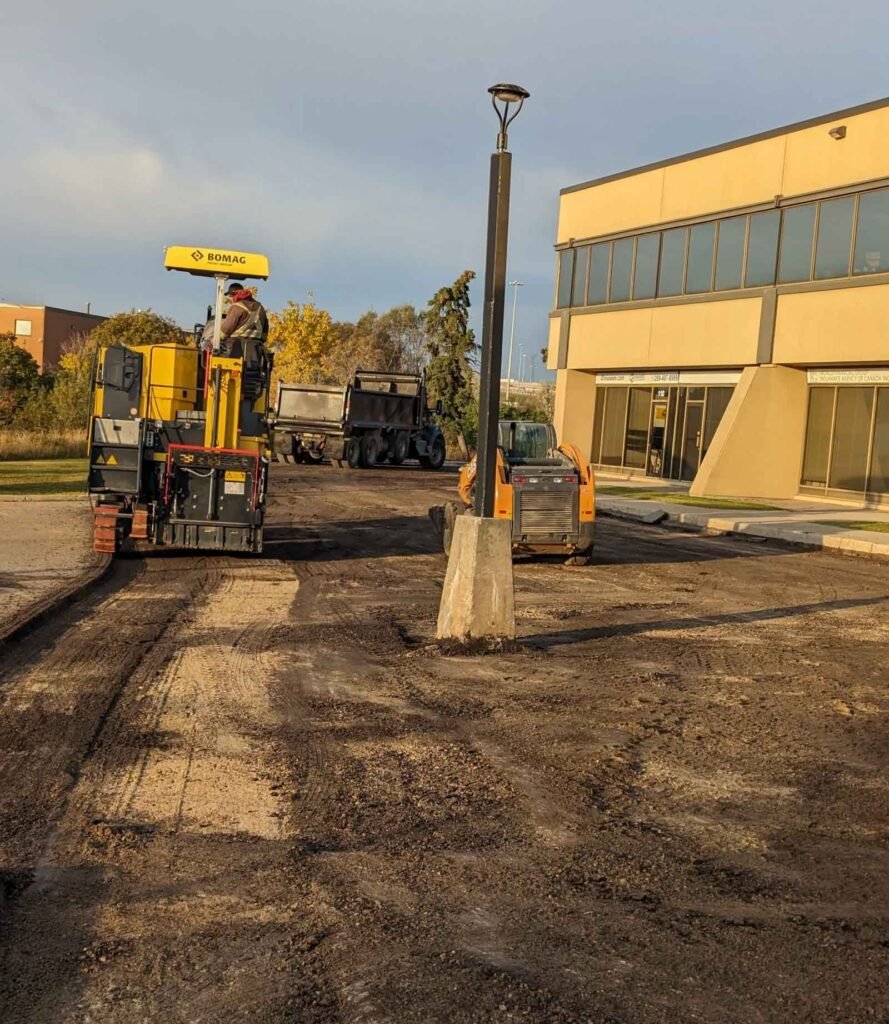 A construction site with heavy machinery, a roller, a truck, and a digger, with a lamp post in the foreground and a building in the background.