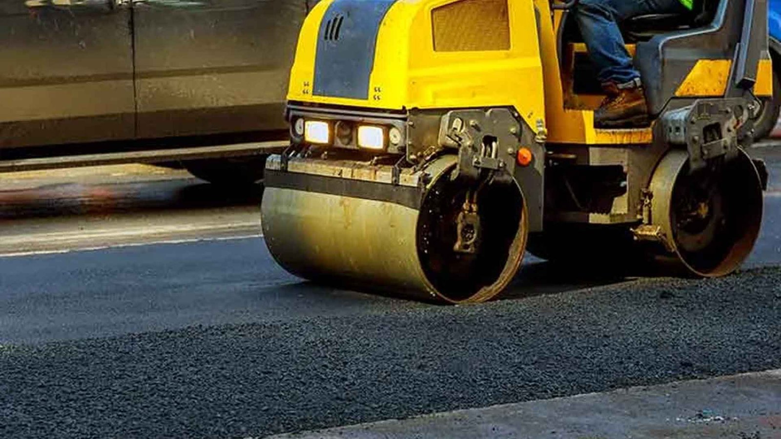 Aman driving yellow roller on paving, wearing safety jacket.