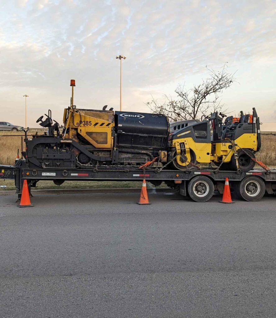 Asphalt paver and road roller machinery on a trailer at a construction site during twilight.