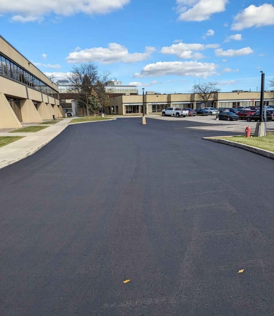 Fresh asphalt on a commercial parking lot with cars and office buildings under a blue sky with clouds.