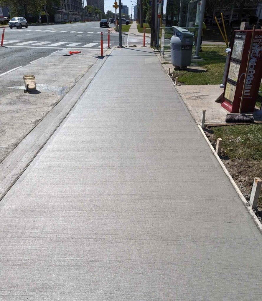 Fresh concrete sidewalk with traffic cones and a city street in the background on a sunny day.