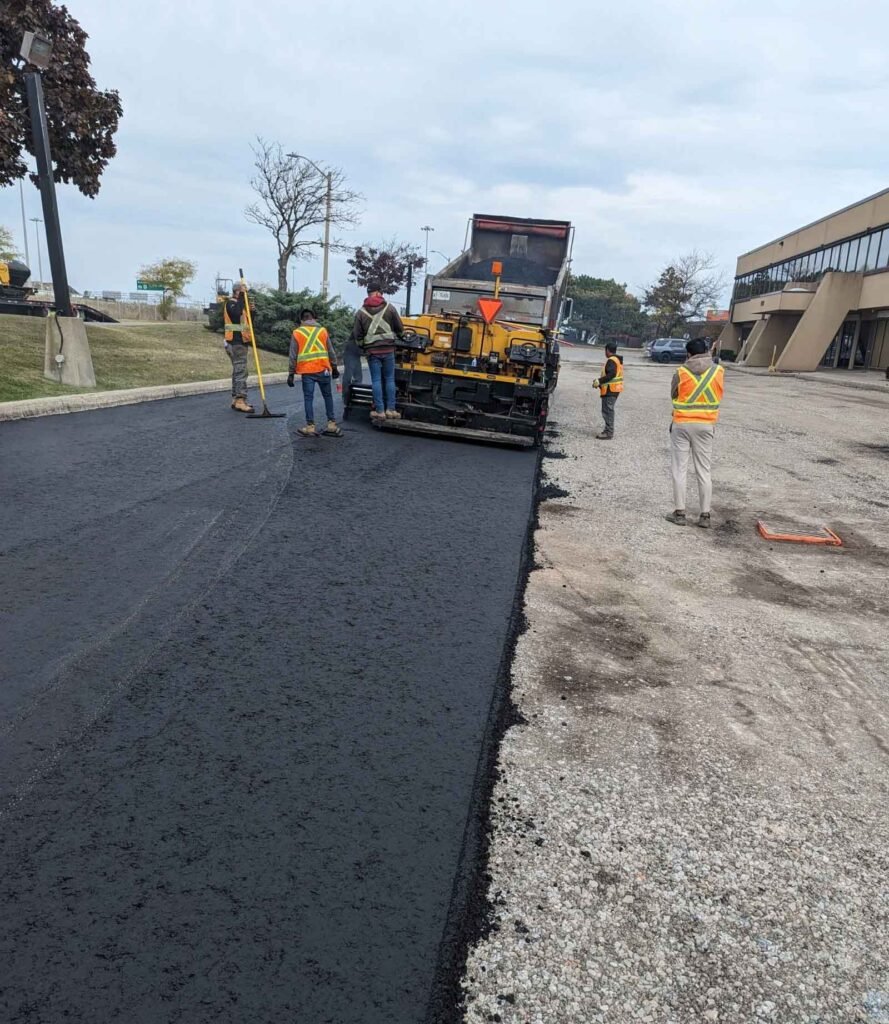 Workers paving a new asphalt road near a commercial building with a paving machine.